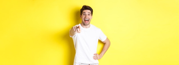 Happy guy pointing finger at camera and laughing check out something standing over yellow background