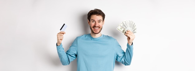 Free photo happy guy smiling and showing plastic credit card with cash choosing between money and contactless p