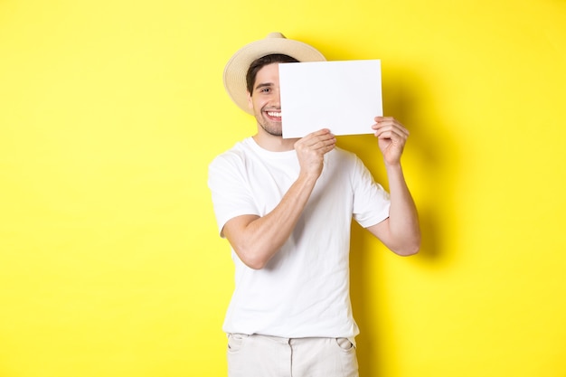 Ragazzo felice in vacanza che mostra un pezzo di carta bianco per il tuo logo, con in mano un cartello vicino al viso e sorridente, in piedi su sfondo giallo
