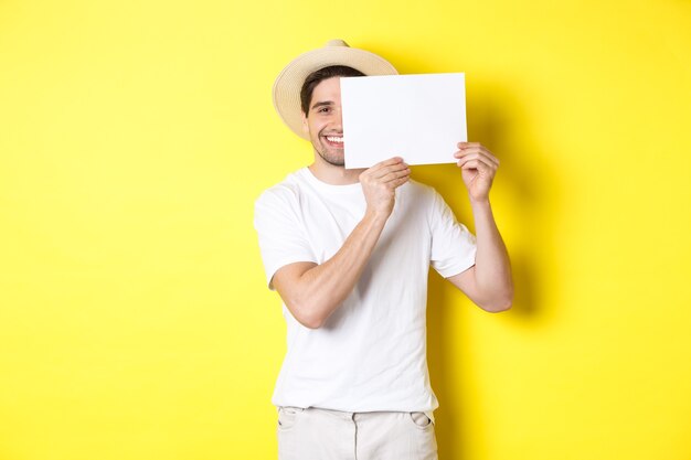 Happy guy on vacation showing blank piece of paper for your logo, holding sign near face and smiling, standing against yellow background