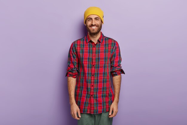 Happy guy with bright smile, stubble, wears yellow hat and checkered shirt, enjoys free time