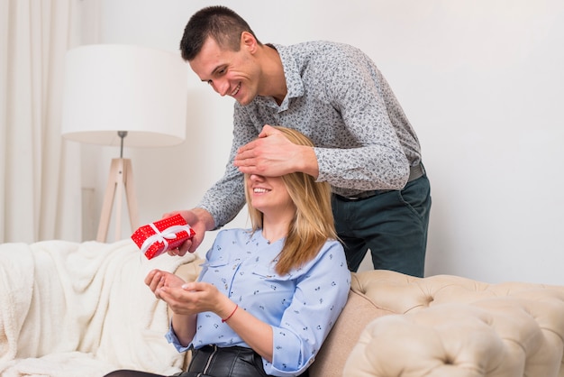 Happy guy with present closing eyes to smiling lady on sofa