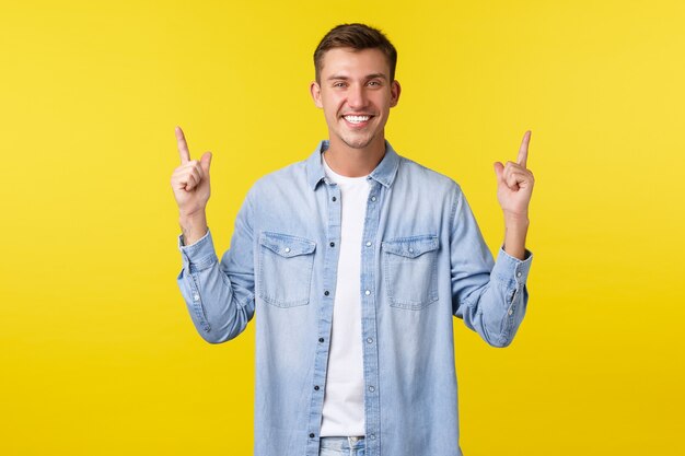 Happy handsome adult man with perfect white smile, pointing fingers up, demonstrating banner, advertising and recommending buy product, special summer discount, standing yellow background.