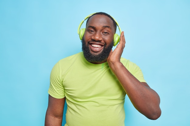 Happy handsome Afro American man with thick beard keeps hand on stereo headphones enjoys perfect sound wears casual green t shirt isolated over blue wall