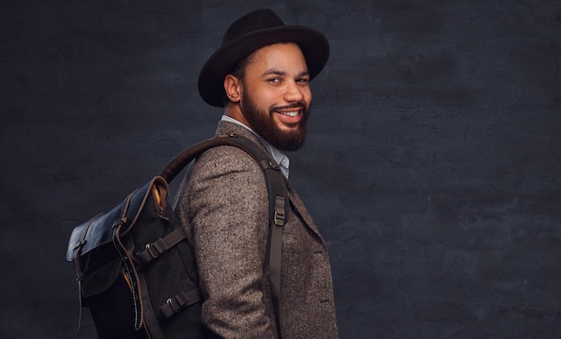 Happy handsome Afro-American traveler in a brown jacket and hat with the backpack, stands in a studio. Isolated on a dark background.