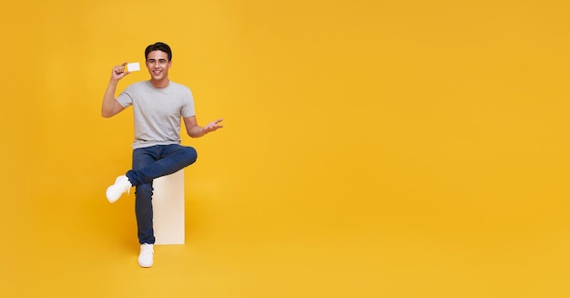 Happy handsome Asian man showing credit card for making payment or paying online business in hand isolated on yellow background