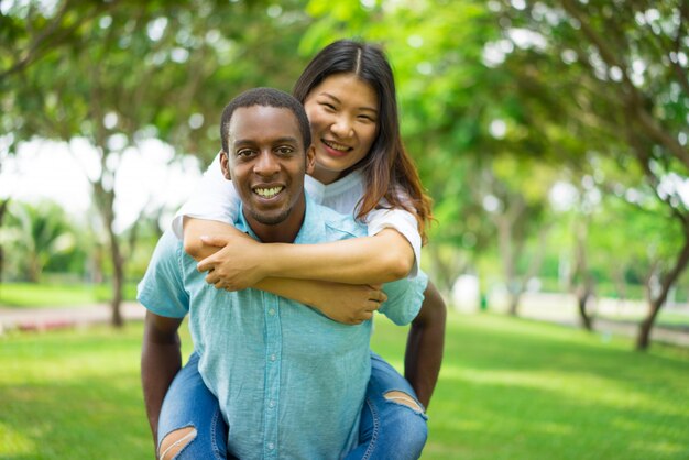 Happy handsome black guy carrying Asian girlfriend on his back while they walk