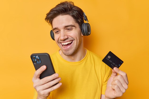 Happy handsome dark haired man types data from credit card in banking app pays easily from home dressed in casual t shirt wears stereo headphones on ears purchases goods in internet poses indoors