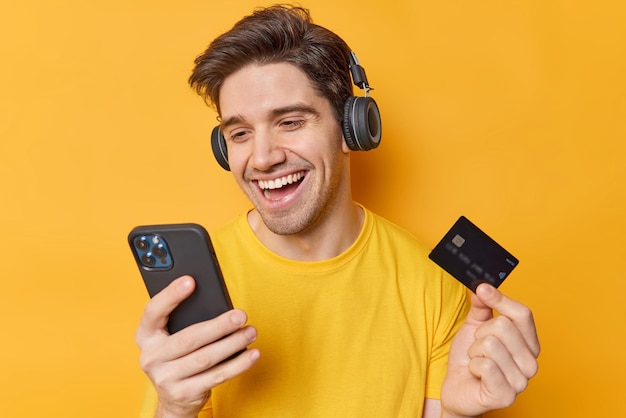 Happy handsome dark haired man types data from credit card in banking app pays easily from home dressed in casual t shirt wears stereo headphones on ears purchases goods in internet poses indoors
