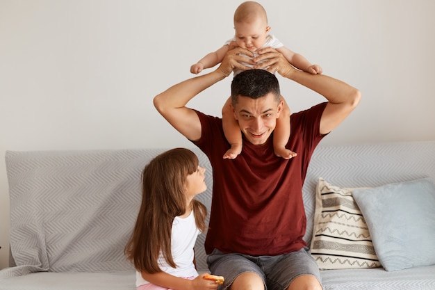 Happy handsome dark haired man wearing casual style clothing posing at home together with his kids, cute infant baby on dad's shoulders, family expressing happiness.