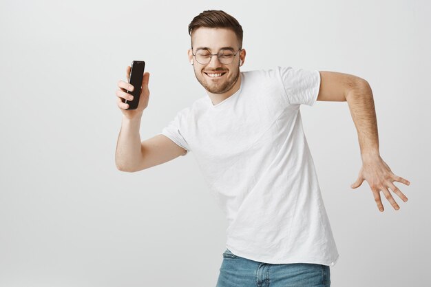 手に携帯電話を持つワイヤレスイヤホンで音楽に合わせて踊るメガネで幸せなハンサムな男