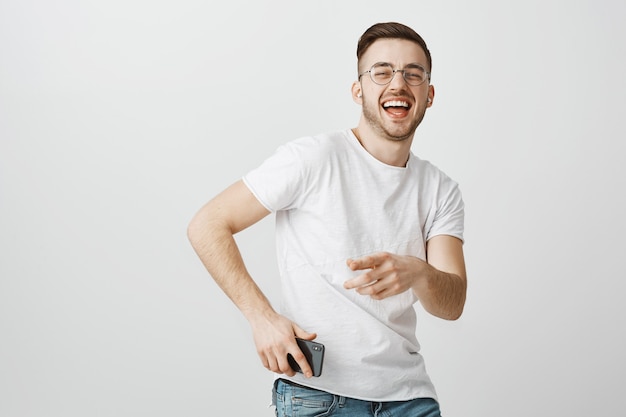 Happy handsome guy in glasses dancing to music in wireless earphones with mobile phone in hand