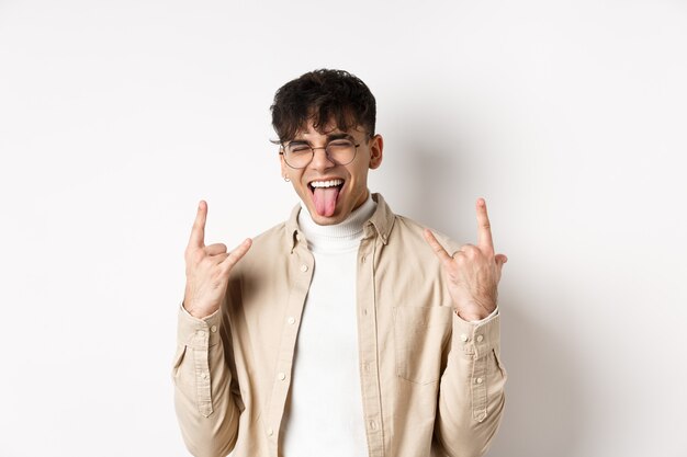 Happy handsome guy showing tongue and rock-on gesture, having fun and feeling upbeat, enjoying something good, standing on white background