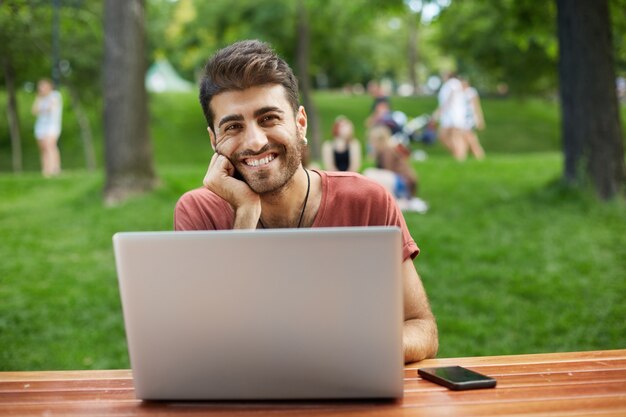 Счастливый красивый мужчина-разработчик, парень-фрилансер сидит в парке с ноутбуком и улыбается