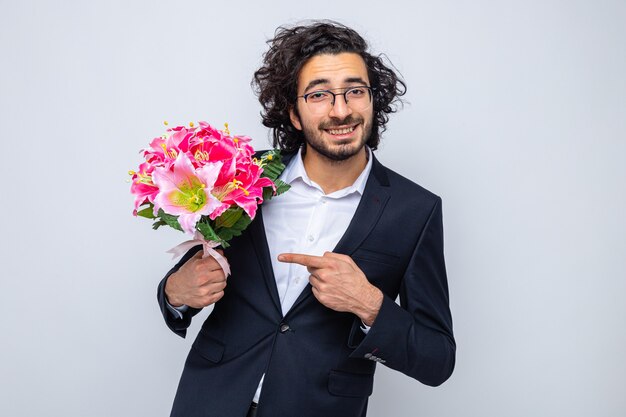 Счастливый красавец в костюме с букетом цветов, указывая на него указательным пальцем, глядя в камеру, весело улыбаясь, празднуя международный женский день 8 марта, стоя на белом фоне