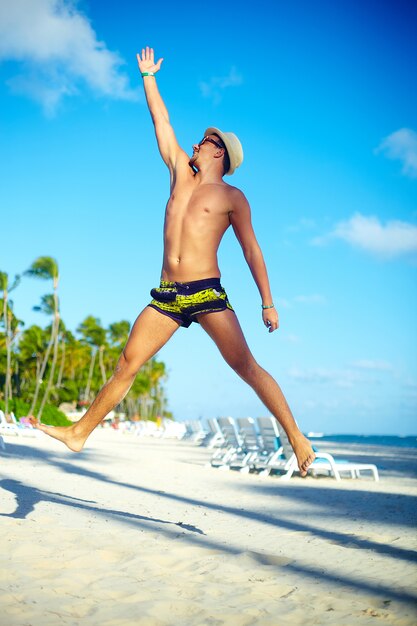 Happy handsome muscled man in sunhat on beach jumping behind blue sky behind blue sky