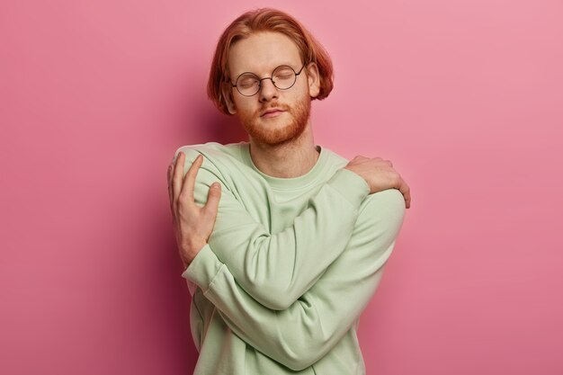 Happy handsome redhead male teenager in spectacles embraces himself, keeps eyes closed, has high self esteem, feels comfort in new bought jumper, isolated on pink wall. Self care concept