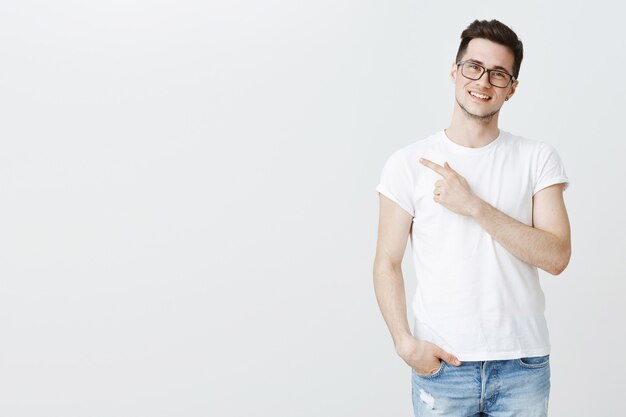 Happy handsome young male student in glasses pointing finger left at copyspace