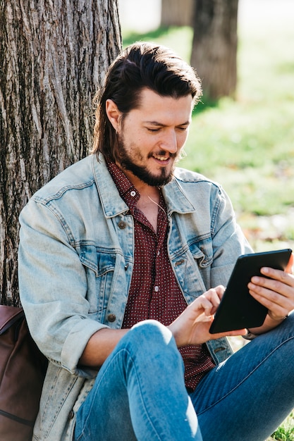 Giovane bello felice che si siede sotto l'albero usando smartphone