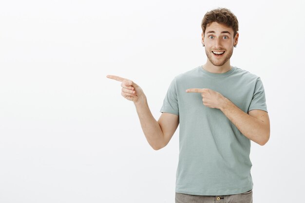 Happy handsome young man with earrings, pointing left with index fingers and smiling joyfully, showing something awesome and interesting