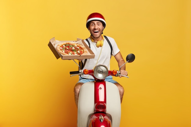 Счастливый трудолюбивый красивый мужчина-водитель на скутере в красном шлеме доставляет пиццу