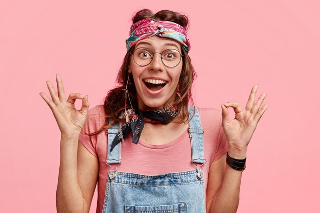 Happy hippie woman makes okay gesture, likes friend`s plan, wears stylish clothes, has joyful facial expression, isolated over pink wall. Hippy woman ready to something