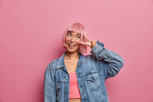 Free photo happy hipster girl has pink hair does victroy gesture, smiles and rejoices success, expresses positive view, has dreamy expression, dressed in fashion clothes. stylish woman makes v sign with fingers