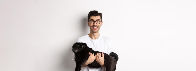 Free photo happy hipster in glasses holding cute black pug and smiling dog owner staring at camera with amazed