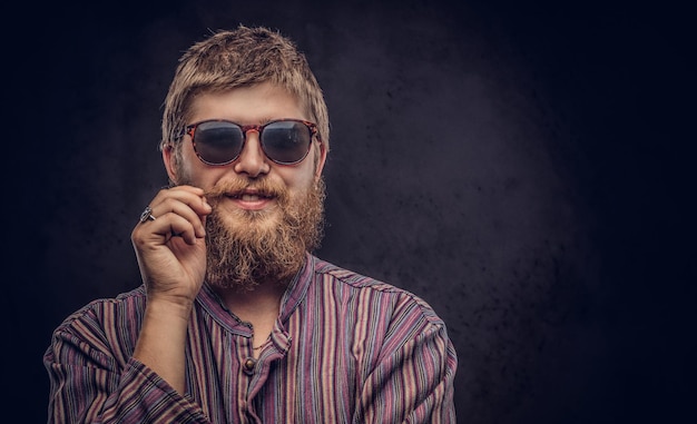 Foto gratuita il ragazzo felice dei pantaloni a vita bassa che indossa occhiali da sole vestito con una camicia antiquata si corregge i baffi. isolato su uno sfondo scuro.