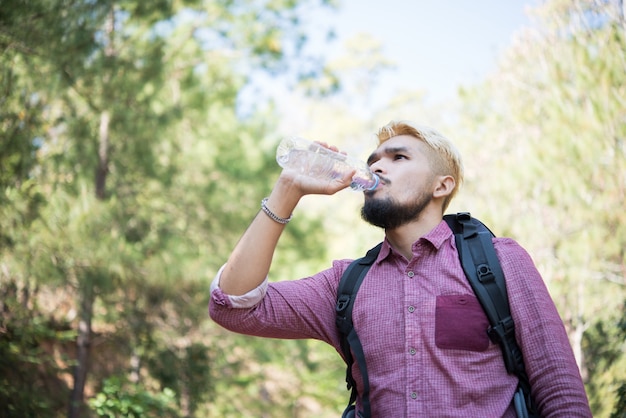 Happy hipster man tourist with backpack drinking water while hiking in nature forest.