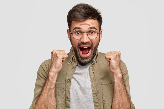 Happy hipster with clenched fists and opened mouth looks joyfully, celebrates victory, wears round spectacles and fashionable shirt