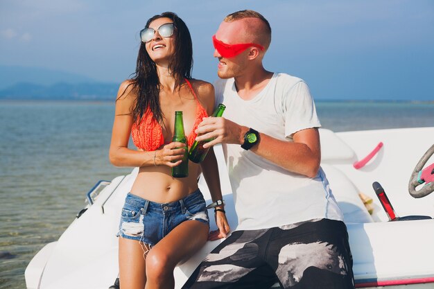 Happy hipster woman and man drinking beer on summer tropical vacation in Thailand traveling on boat in sea, party on beach, people having fun together, positive emotions