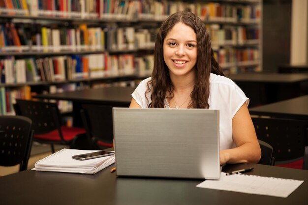 Happy Hispanic university student using a laptop for homework in the school library