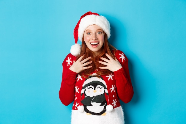 즐거운 휴일과 크리스마스 컨셉입니다. 예상치 못한 선물을 받고 놀란 빨간 머리 소녀는 깜짝 놀라 숨을 헐떡이며 유쾌한 불신으로 쳐다보며 파란색 배경에 산타 모자를 쓰고 서 있습니다.