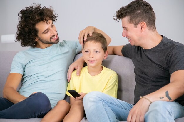 Happy homosexual dads and son watching TV show together at home, sitting on couch in living room, smiling and hugging boy. Family and home entertainment concept