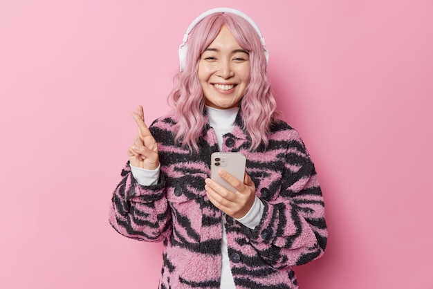 Happy hopeful teenage girl with pink hair crosses fingers for good luck makes wish uses mobile phone and headphones for listening playlist dressed in fashionable coat. Modern technologies concept