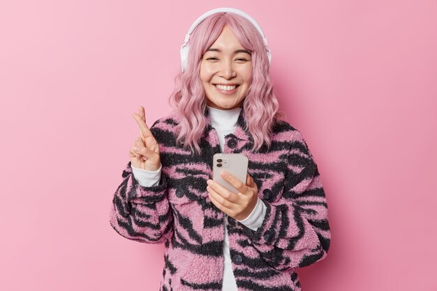 Happy hopeful teenage girl with pink hair crosses fingers for good luck makes wish uses mobile phone and headphones for listening playlist dressed in fashionable coat. Modern technologies concept