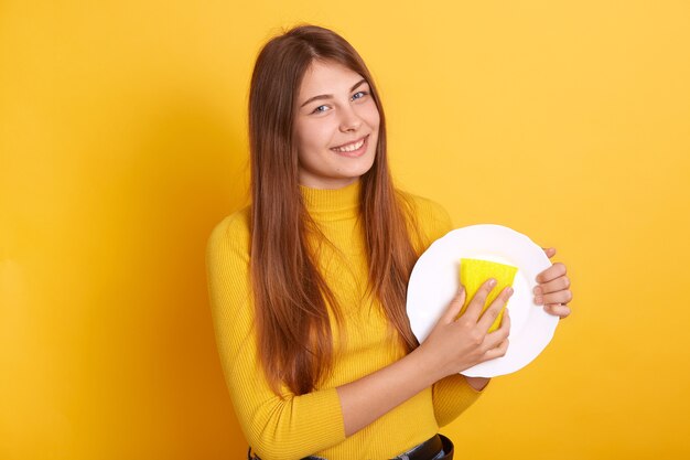 Happy housewife demonstrating washing process, holding white plate and sponge in hands and , wearing casual attire