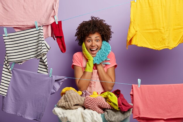 Happy housewoman hangs clean laundry on clothes line, does washing at home, busy with household duties holds mop, wears t shirt and rubber gloves, dries clothes, pegs out washing, smiles broadly