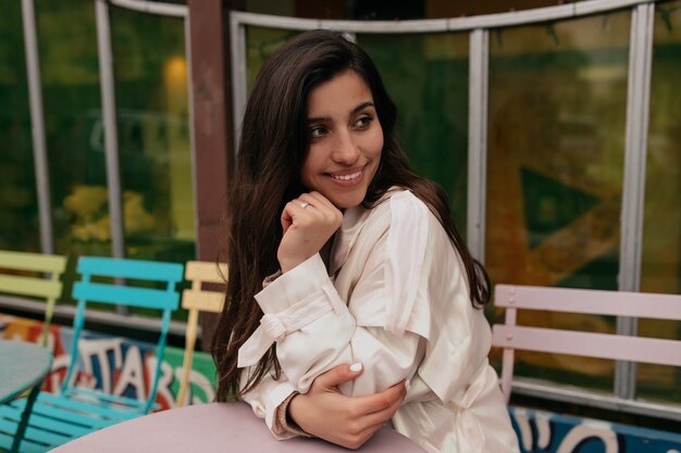 Happy incredible woman with long hair wearing beige coat smiling and waiting for her order in open air cafe