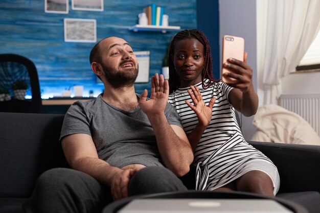 Happy interracial married couple waving at modern phone while in videocall. Cheerful diverse people talking to their family through online videocall while sitting on sofa in living room.