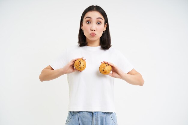 幸せな日本人女性は、白い背景の上に立ってペストリーを食べて胸にカップケーキを示しています