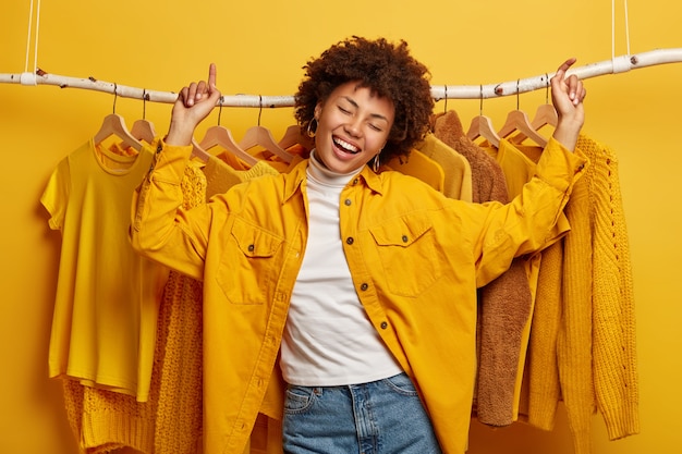Happy joyful Afro woman dances with triumph against clothes rack, prefers outfits of yellow colour, wears fashionable jacket and jeans, moves actively near home wardrobe.