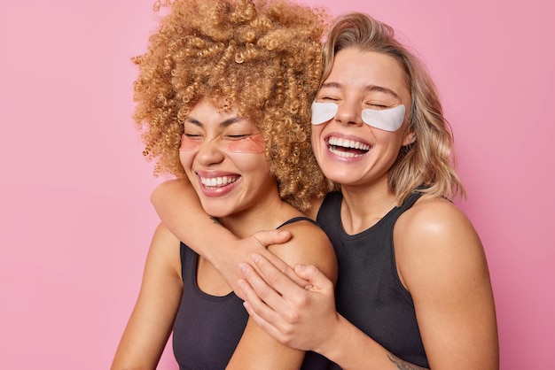 Free photo happy joyful female models embrace and laugh gladfully keep eyes closed have fun undergo beauty treatments apply patches have healthy skin isolated over pink background friendship emotions concept