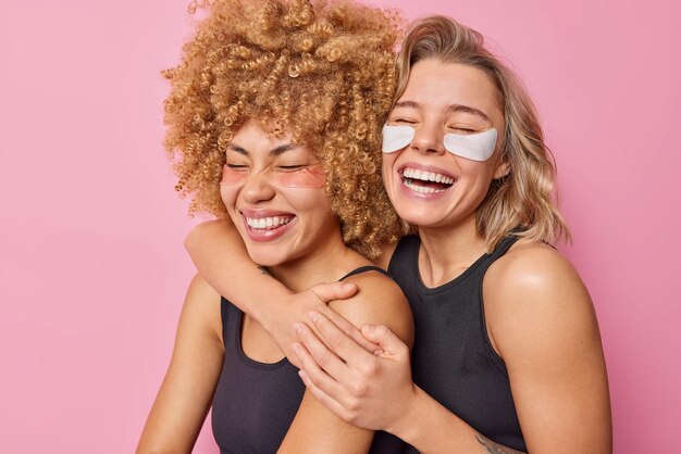 Happy joyful female models embrace and laugh gladfully keep eyes closed have fun undergo beauty treatments apply patches have healthy skin isolated over pink background Friendship emotions concept