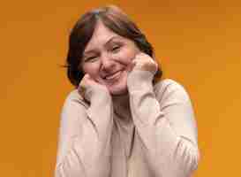 Free photo happy and joyful middle aged woman in beige turtleneck  with hands on her cheeks standing over orange wall