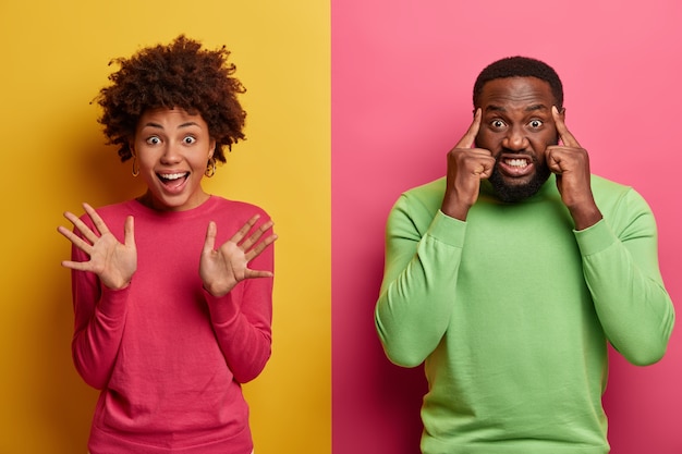 Happy joyful woman raises palms, waves hands in hi, makes friendly gesture, intense bearded man touches temples, tries to concentrate, clenches teeth, has headache, stand against two colored wall