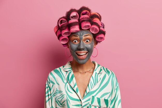 Happy joyful woman visits hairdressing and spa salon, makes perfect hairstyle and applies clay face mask, wears pyjama, has surpised expression, isolated on pink . Female going on date