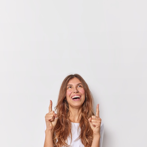 Happy joyful woman with long hair indicates index fingers overhead shows something above smiles broadly demonstrates blank copy space for advertising content shows promotion over white wall.