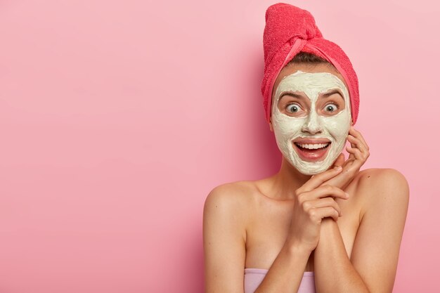 Happy joyful young woman applies cream mask wonderful for any skin type, wears red towel on head, nourishes skin, looks positively